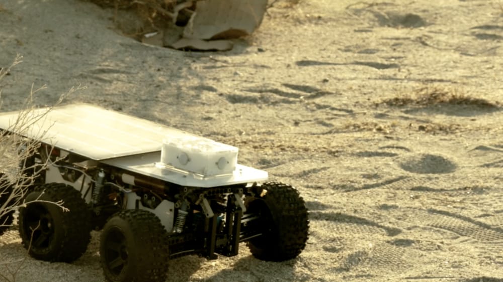 A small robotic rover with large wheels on a sandy terrain in sunlight.