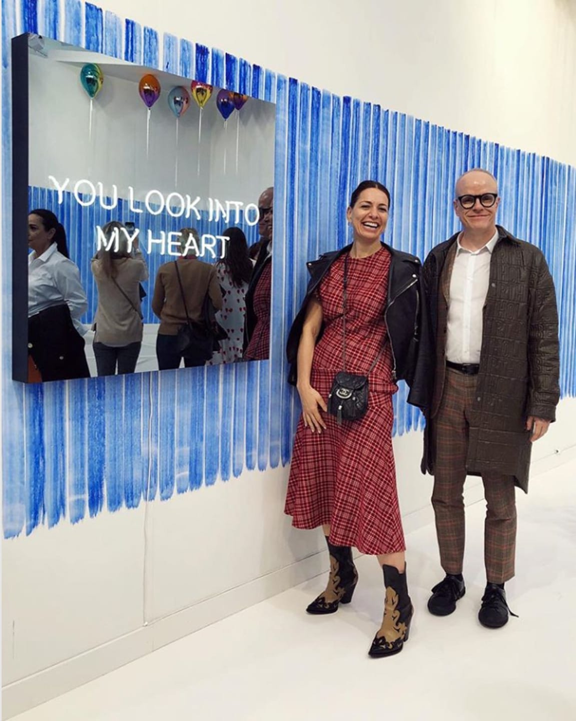 Two people smiling next to an art piece with balloons and the phrase "You look into my heart" on a blue-striped wall.