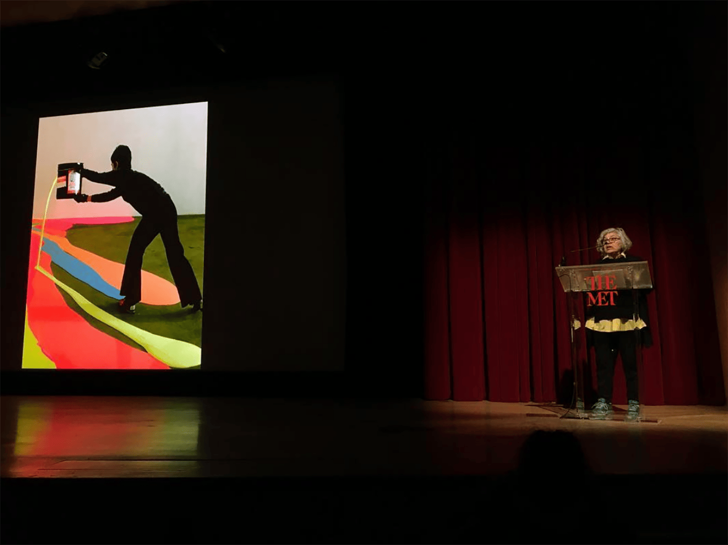 Woman speaking at podium beside large colorful artwork on stage in dimly lit theater.