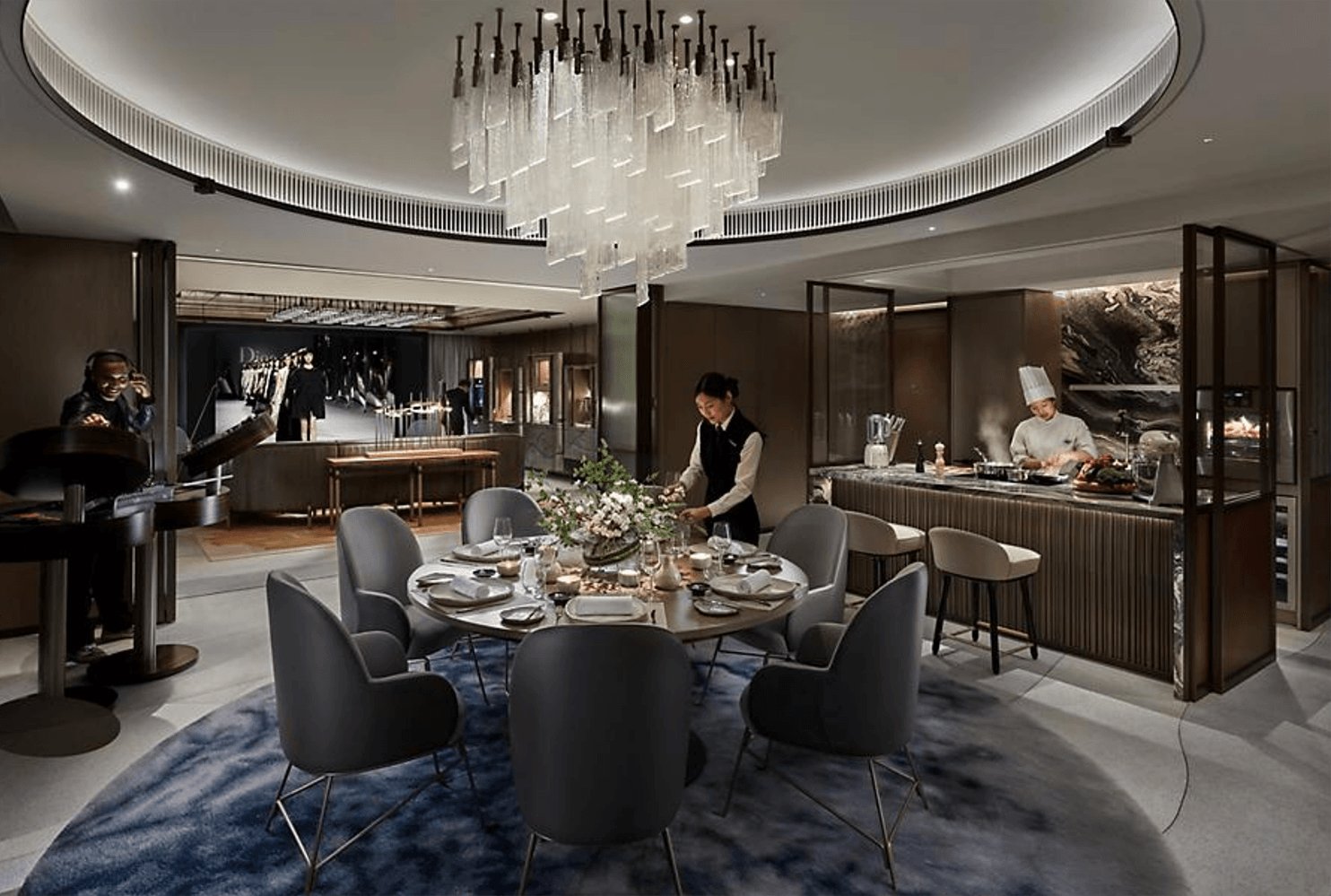 Elegant restaurant interior with dim lighting, round table set for dining, modern chandelier, and chefs preparing food.