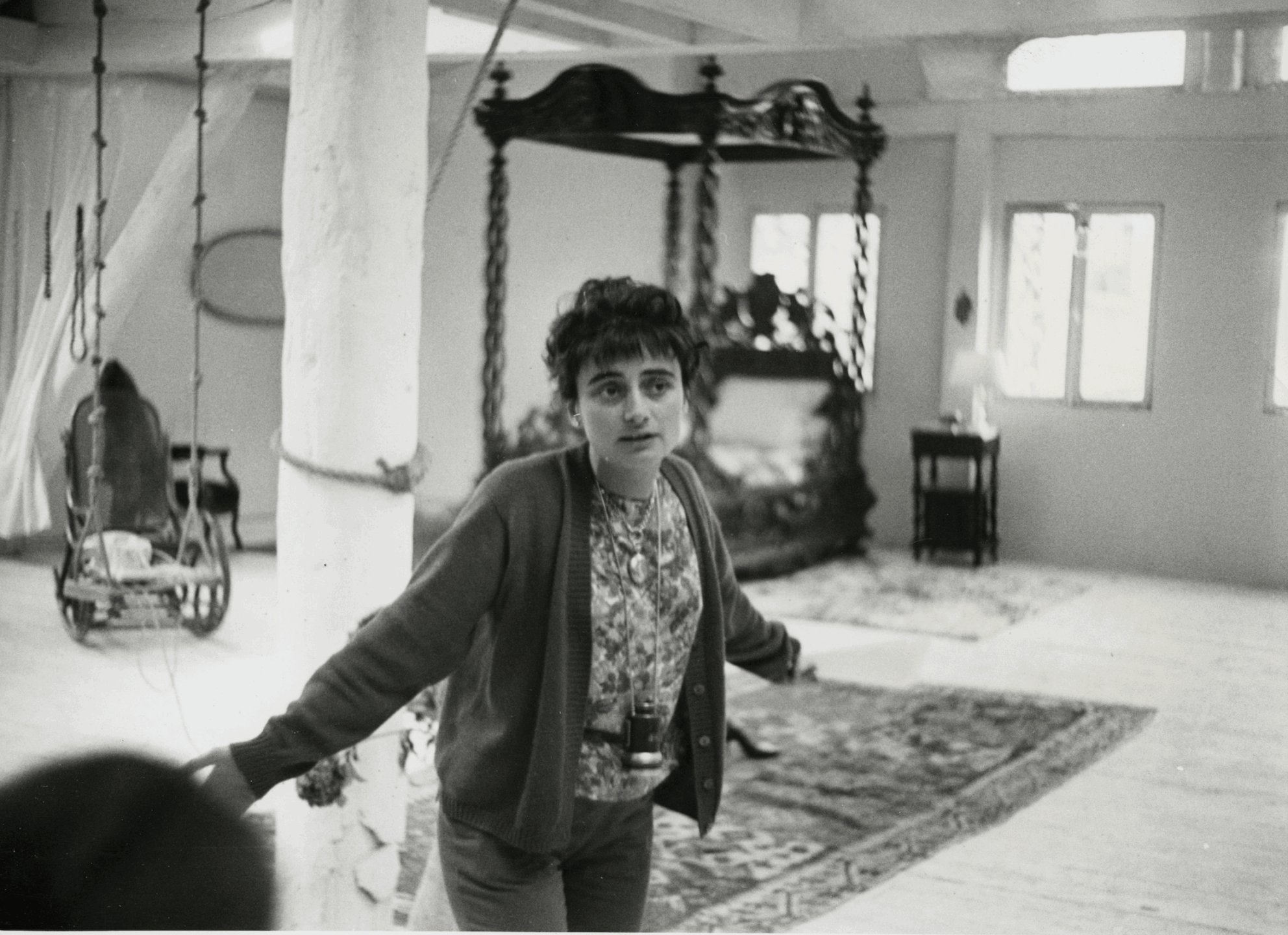 Person standing in a room with a swing and canopy bed, wearing a patterned shirt and cardigan, looking towards the camera.
