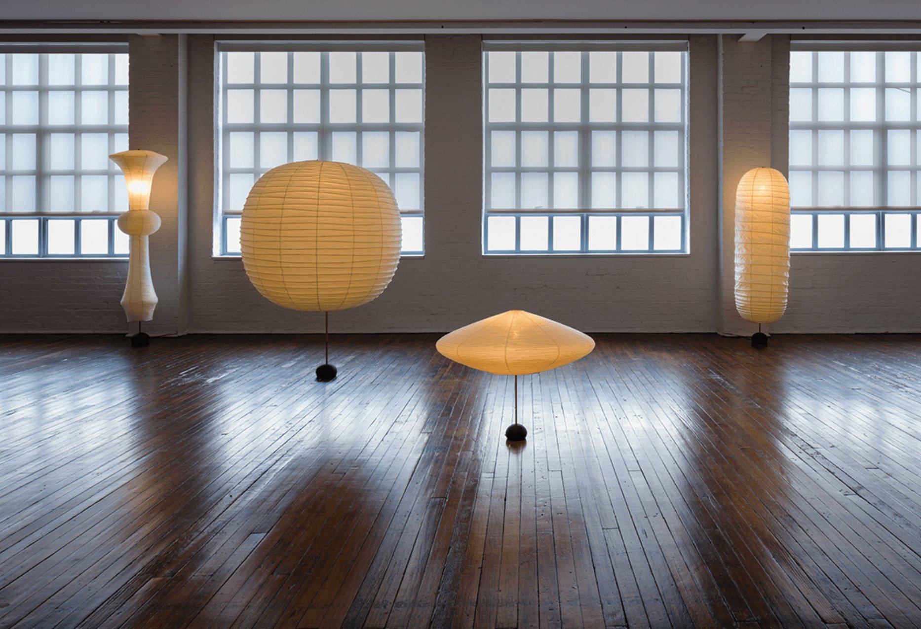 Four modern, illuminated paper lanterns of various shapes displayed in a spacious wood-floored room with large grid windows.