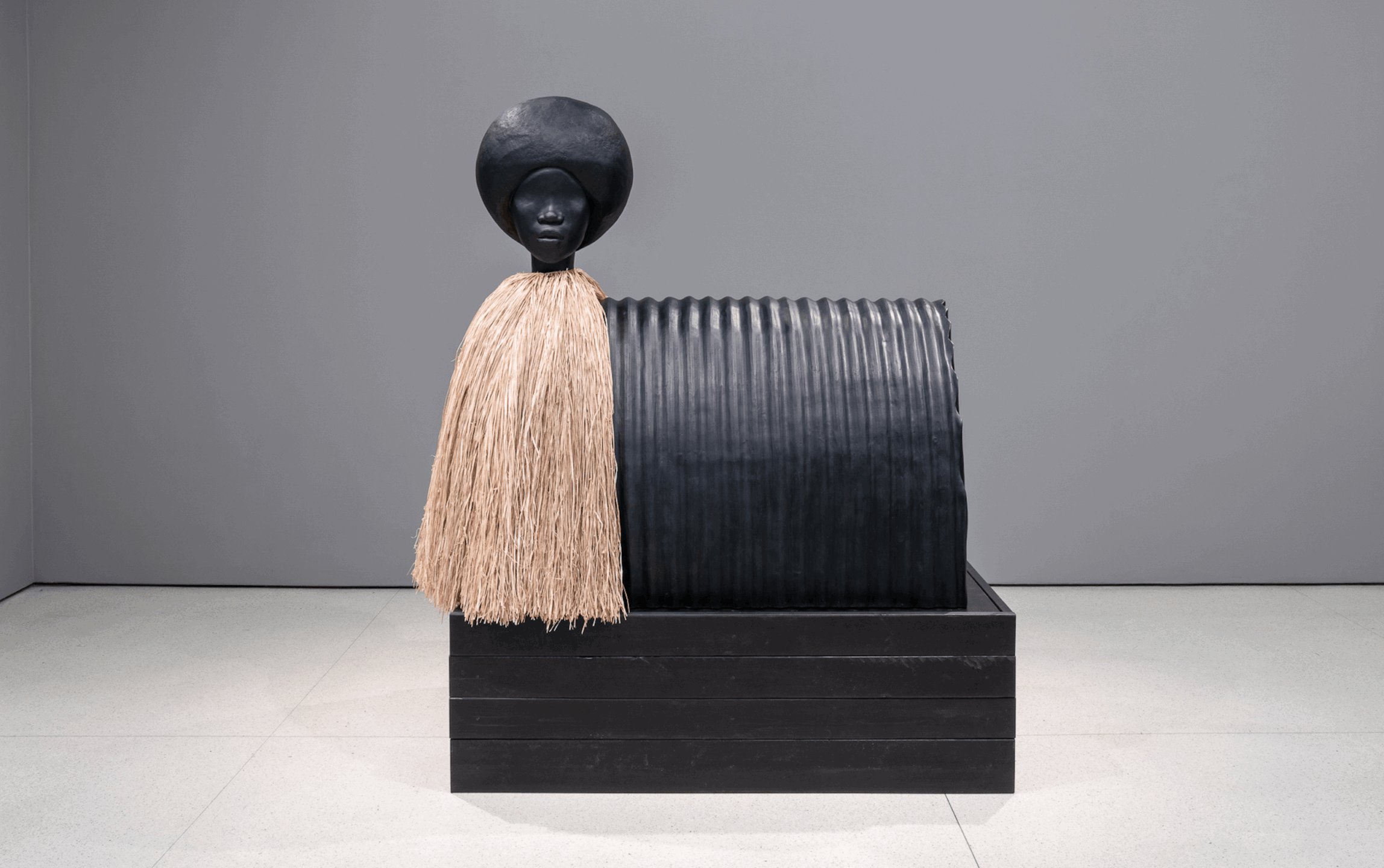 Sculpture of a figure with a black rounded hat, textured cylindrical body, and beige skirt, displayed on a black platform.