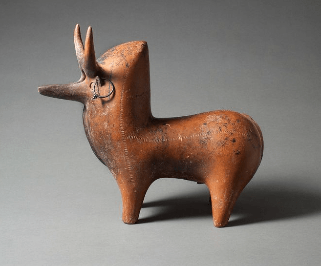 Ancient terracotta sculpture of a stylized animal with long horns on a gray background.