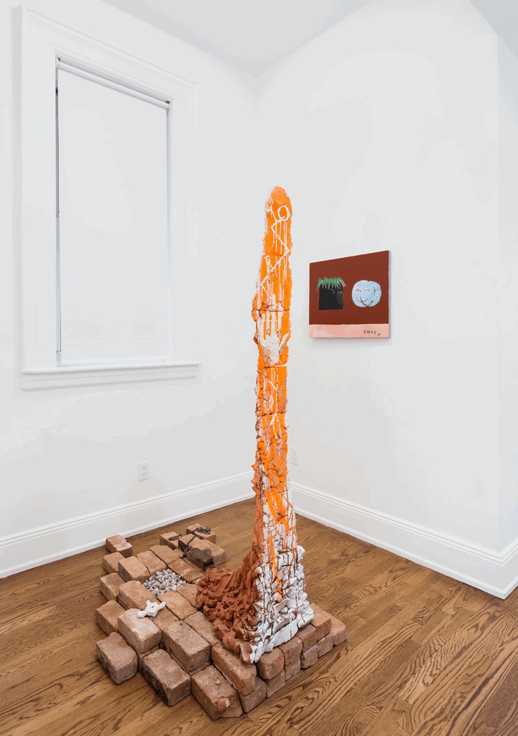 Colorful abstract sculpture stands tall on stacked bricks in a white gallery with paintings on the wall.
