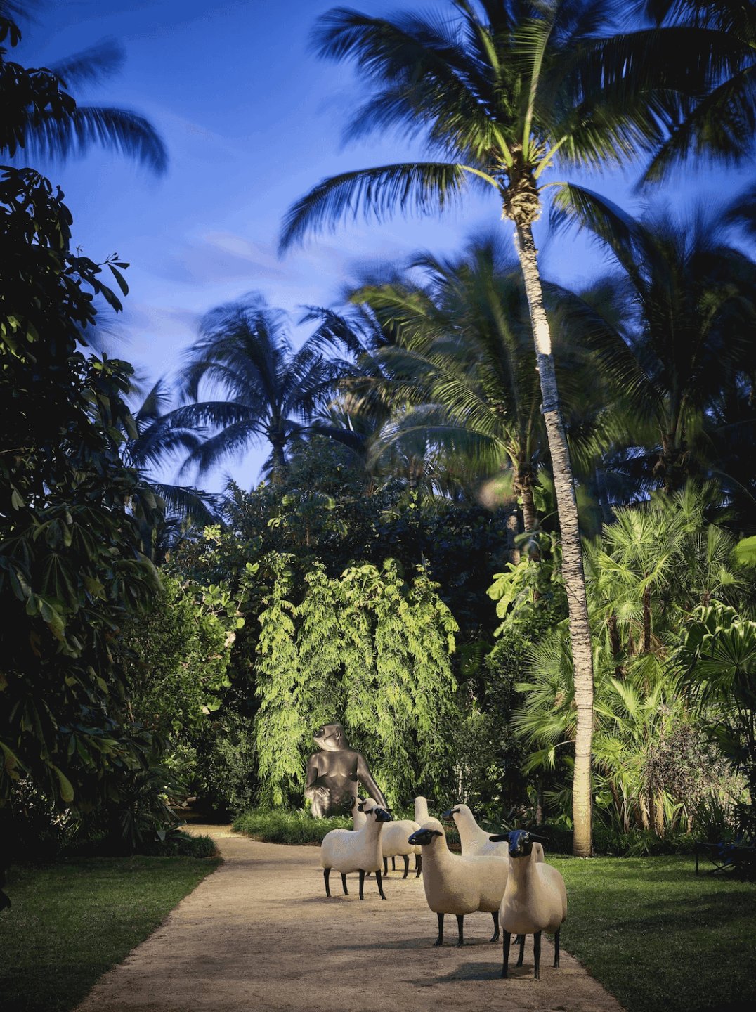 Pathway in tropical garden with palm trees and sheep sculptures under a clear blue sky.