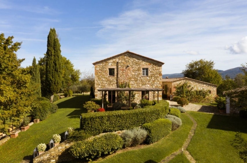 Stone farmhouse with lush garden and trees, under a clear blue sky.