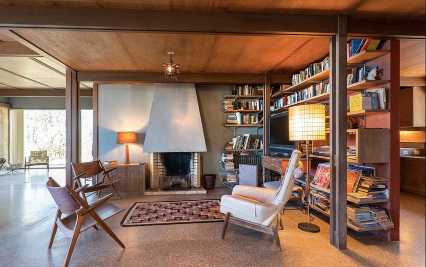 Mid-century modern living room with wooden furniture, bookshelves, a fireplace, and large windows providing natural light.
