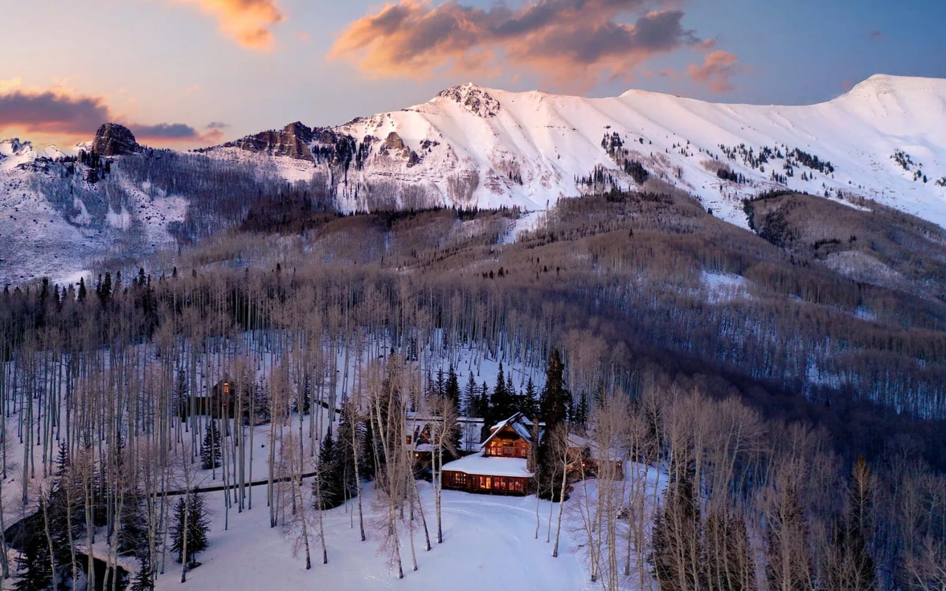 Mountain cabin surrounded by snow-covered trees at sunset, with a backdrop of majestic snowy peaks and a colorful sky.