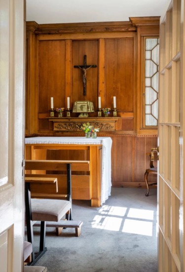 Small wooden chapel with altar, crucifix, lit candles, and a chair in a sunlit room through an open door.