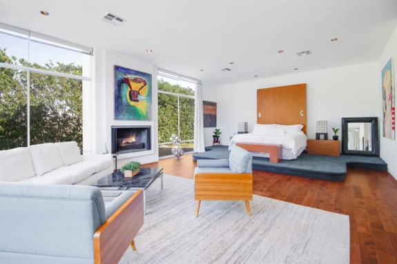 Modern bedroom with a fireplace, large windows, white sofa, blue chair, and hardwood floors, featuring colorful artwork.