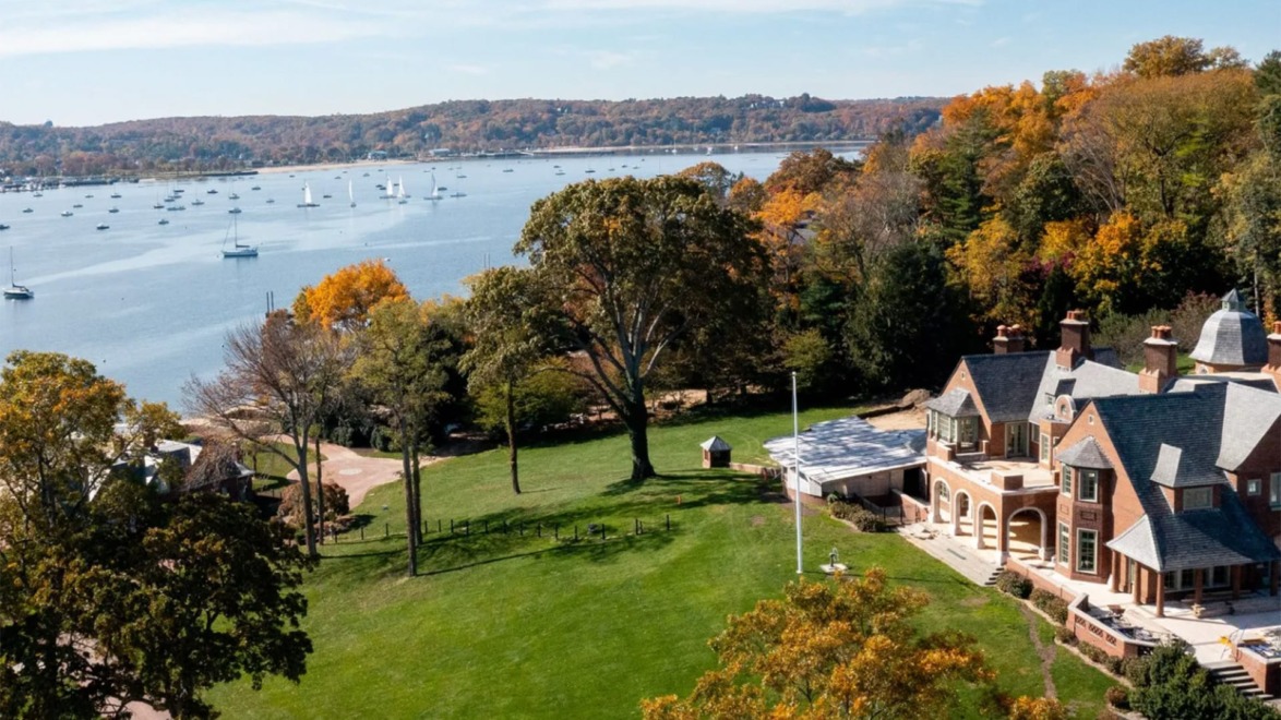 Aerial view of a large lakeside estate with autumn trees, sailboats on the water, and a clear blue sky.