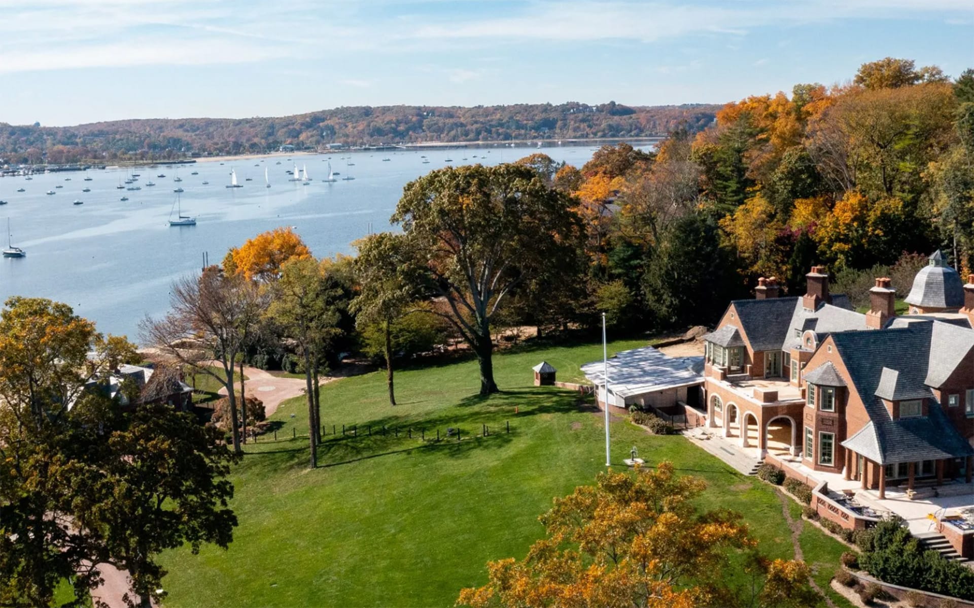 Aerial view of a large lakeside estate with autumn trees, sailboats on the water, and a clear blue sky.
