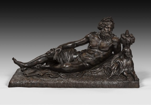 Bronze statue of a reclining bearded man holding grapes, beside a sphinx figure, on a textured base.