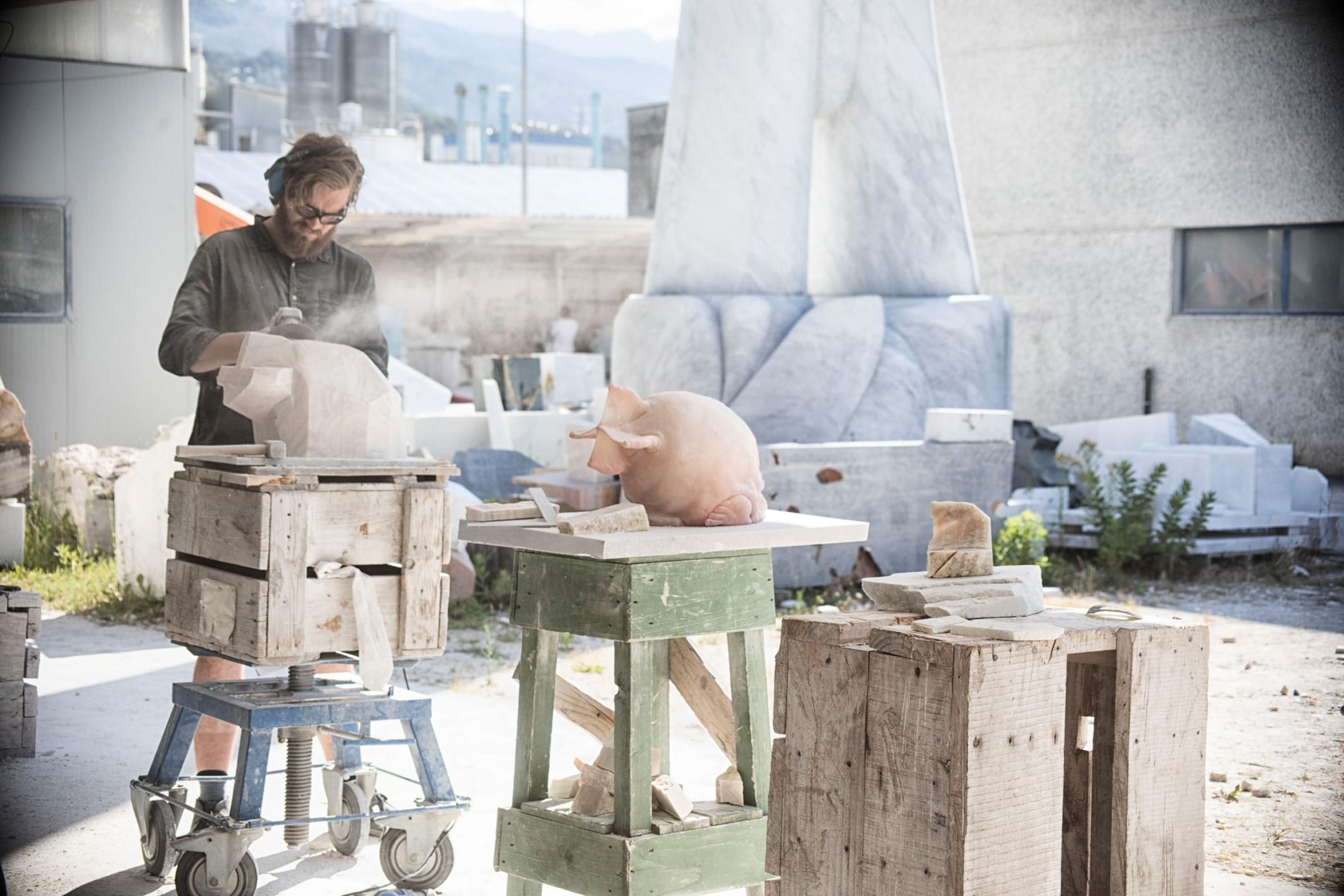 Artist sculpting stone in outdoor workshop with large stone blocks and tools surrounding him.