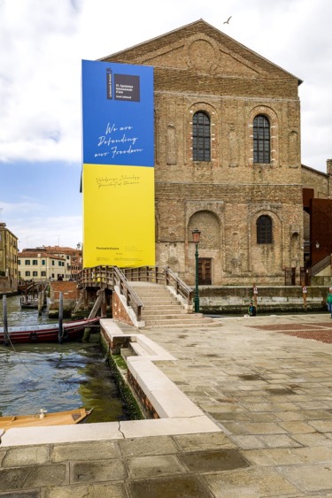 Brick building with large blue and yellow banner displaying text, surrounded by canal water and stone walkway under a cloudy sky.