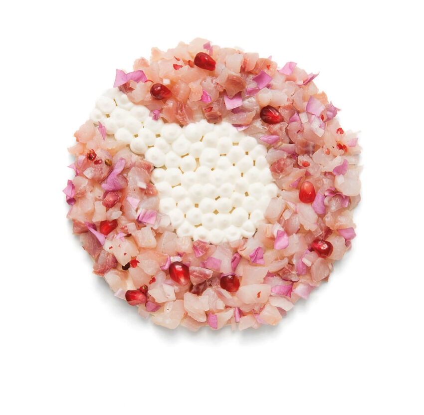 A decorative round dish with flower petals, diced ingredients, white dots, and pomegranate seeds arranged artistically.
