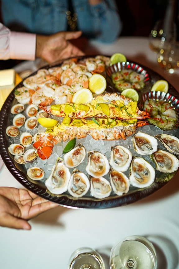 Seafood platter with oysters, shrimp, and lobster garnished with lemon slices, served on ice at a restaurant table.