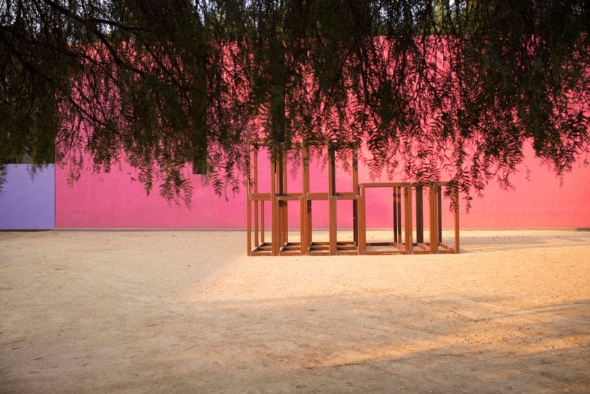 Wooden sculpture under tree branches, set against a pink wall in a sunlit, open outdoor space.