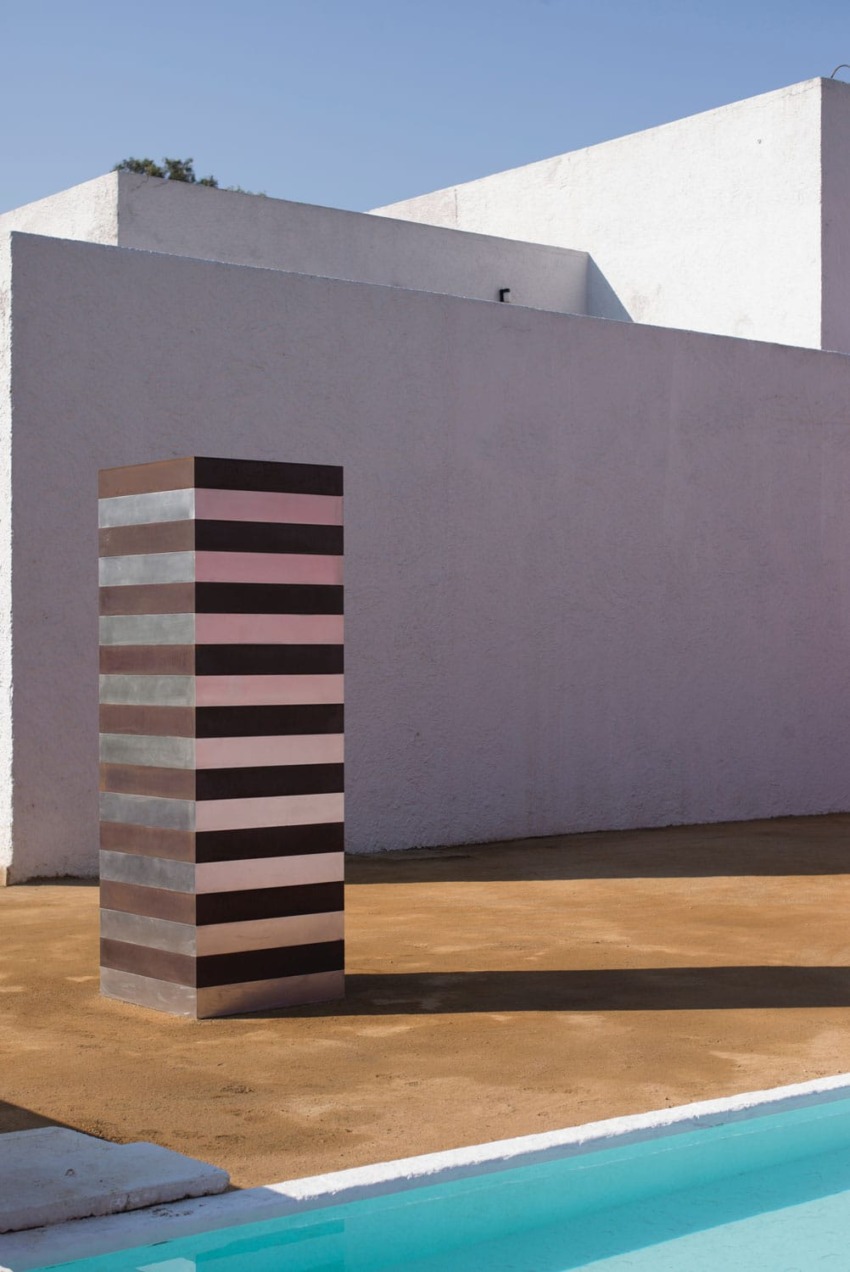 Modern striped sculpture standing on a patio against a white wall with clear blue sky in the background.