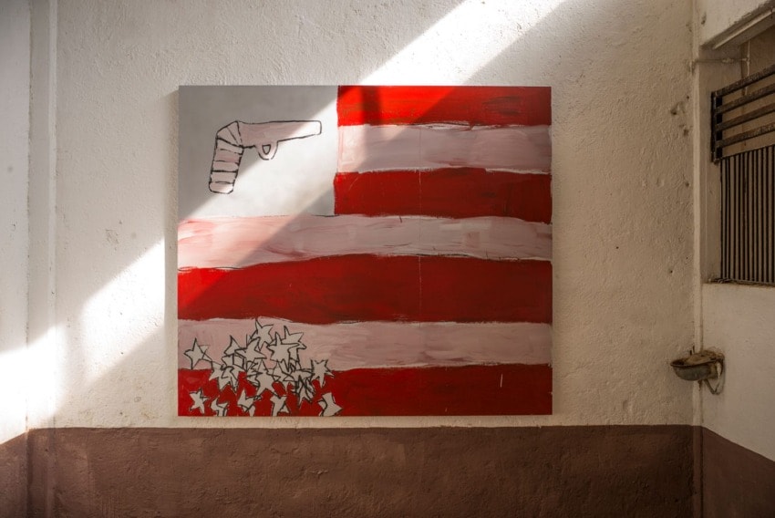 Abstract painting of a gun and stars on a red and white striped background. Sunlight casts a diagonal shadow across the artwork.