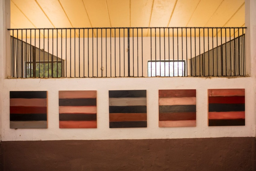 Five abstract paintings with horizontal stripes in various colors hang on a wall below a yellow ceiling and metal railings.