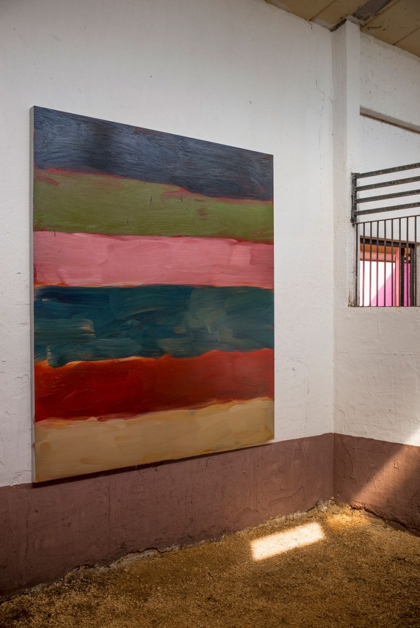 Abstract painting with horizontal stripes in various colors hung on a white and brown wall inside an industrial space.