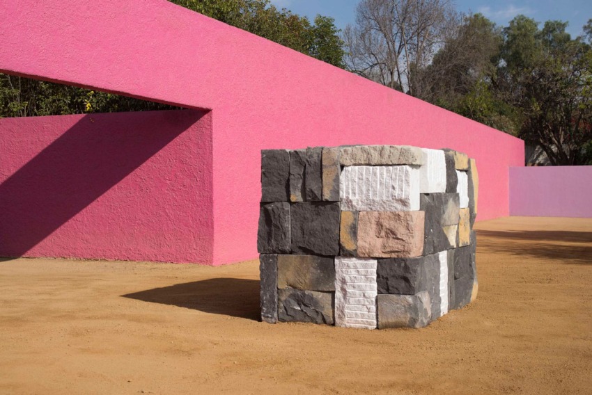 Multicolored stone sculpture in front of vibrant pink walls and sandy ground in an outdoor setting.