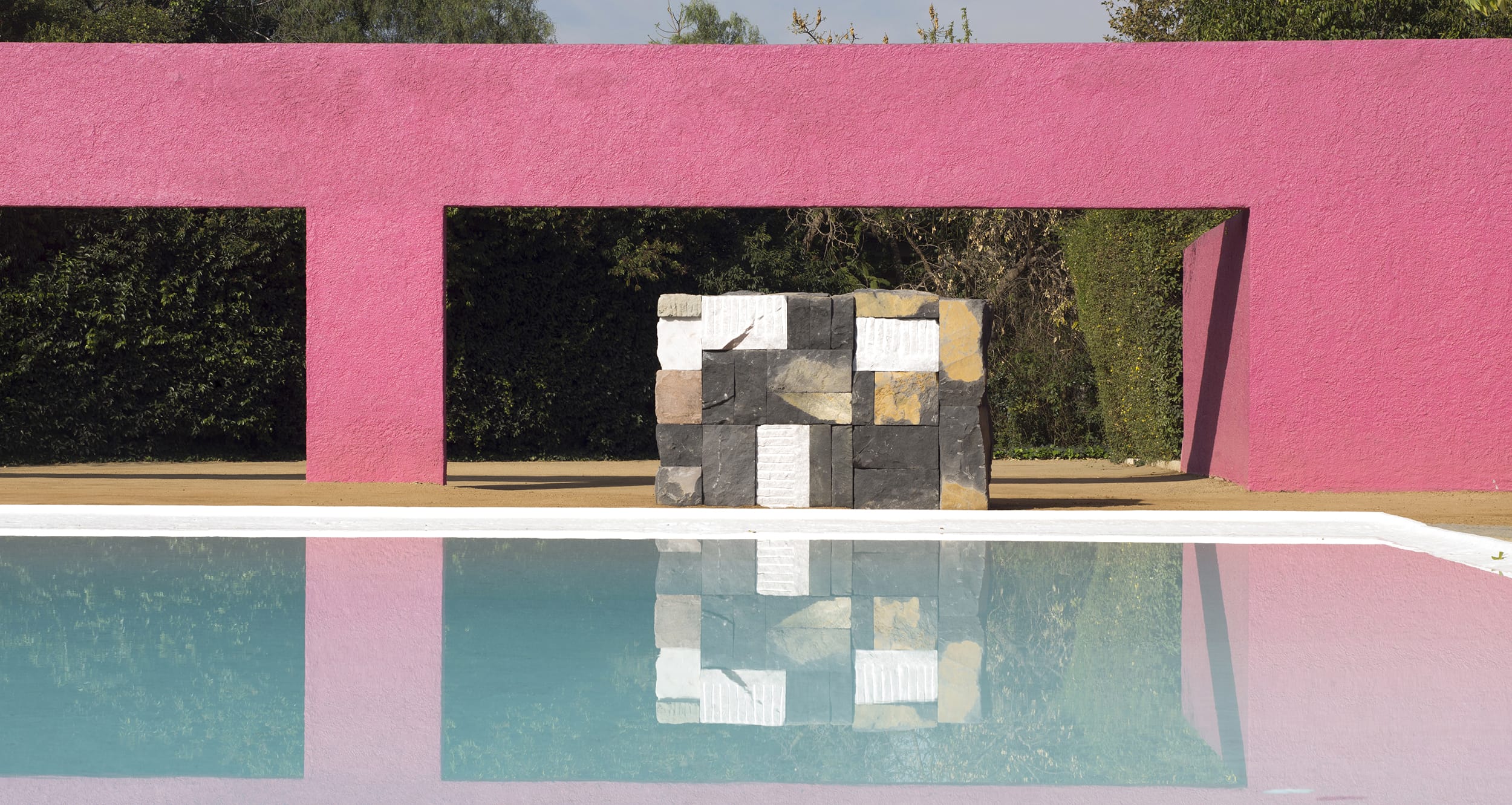Modern art sculpture with geometric shapes reflected in a tranquil pool, set against a bright pink wall and greenery.