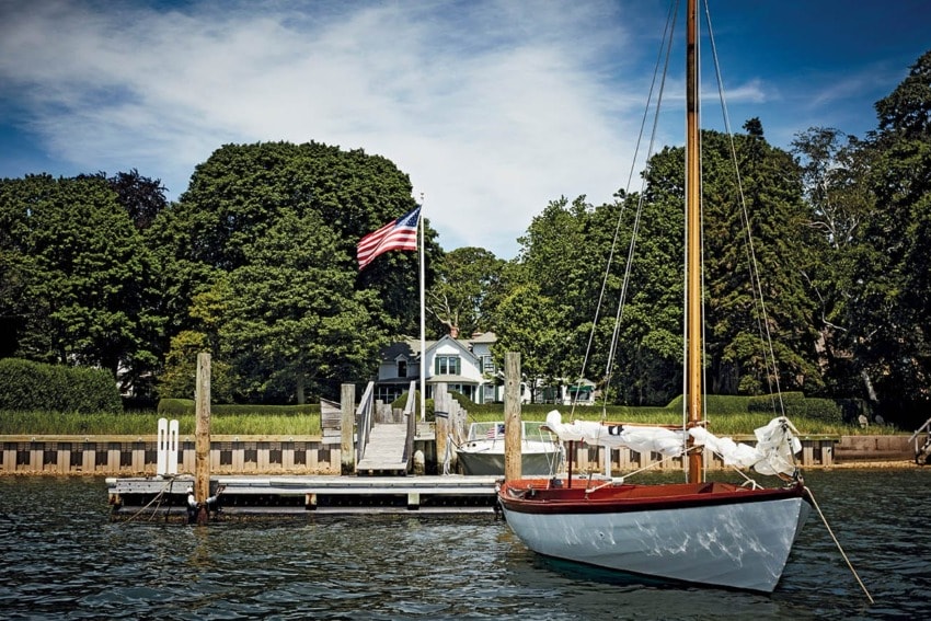 A sailboat docked by a pier with an American flag waving, surrounded by lush green trees and a house in the background.