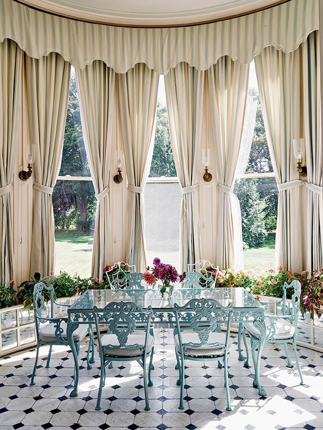 Elegantly decorated sunlit room with tall windows, vintage blue chairs around a glass table, and patterned floor tiles.