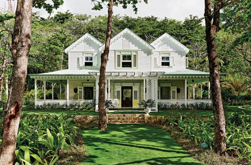 White Victorian-style house with a wraparound porch, set in a lush garden with tall trees in the background.