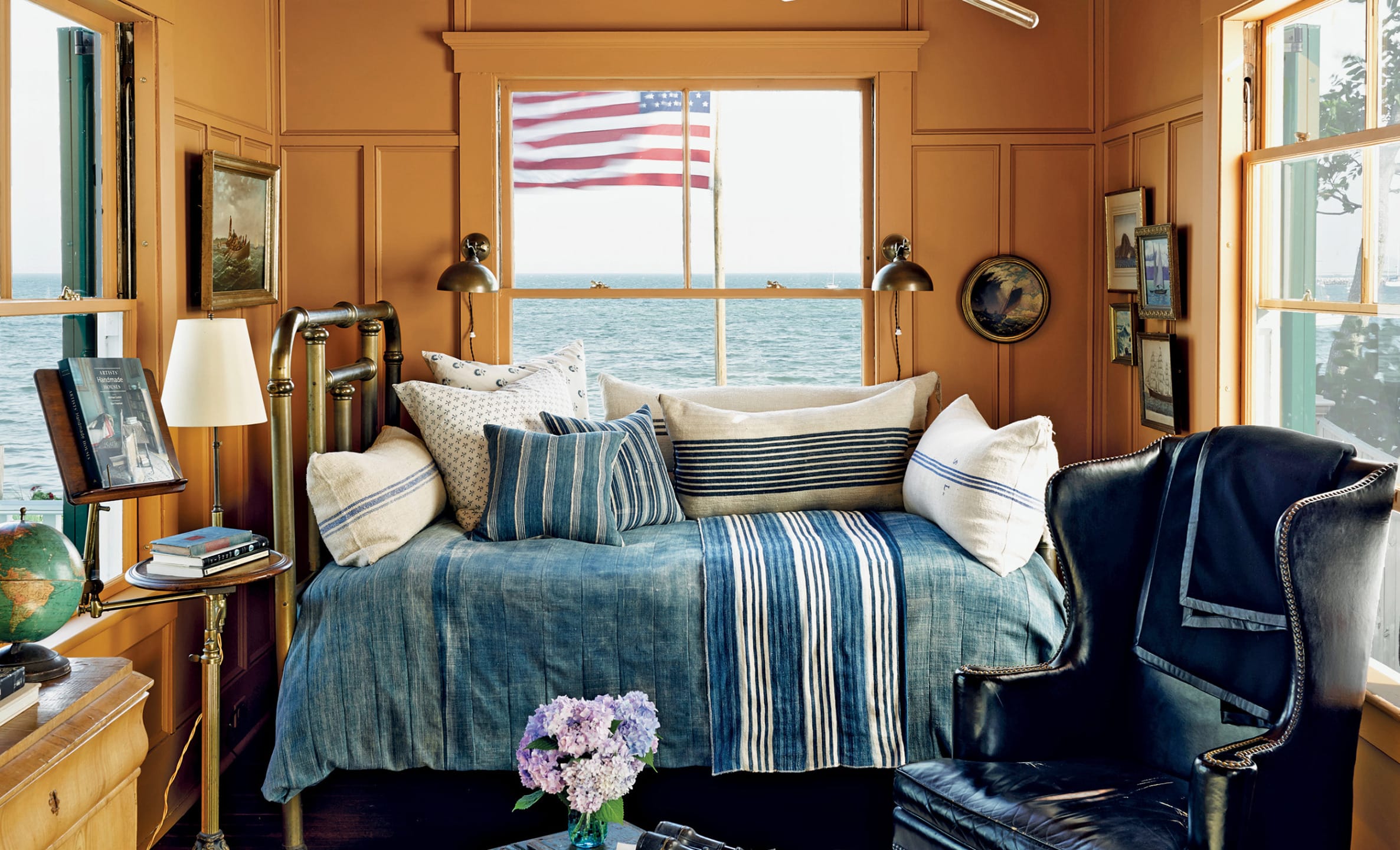 Cozy seaside room with a daybed, striped pillows, American flag out the window, and an ocean view.