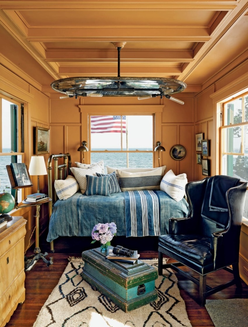 Cozy room with nautical decor, a blue bed, armchair, vintage suitcase, and a window view of the ocean and American flag.