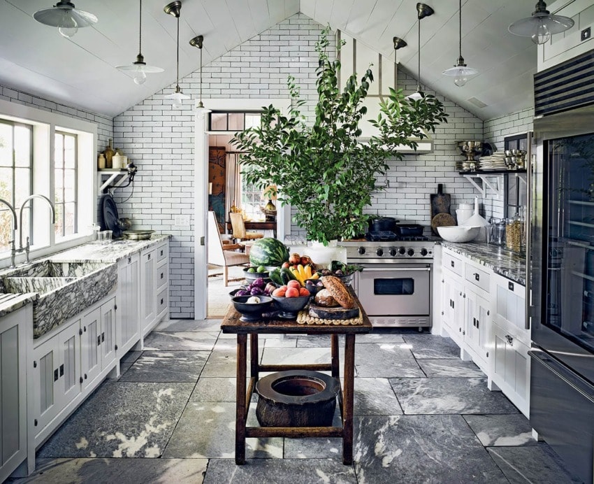 Rustic kitchen with marble floor, white cabinetry, central wooden table with fresh produce, and a large indoor plant.
