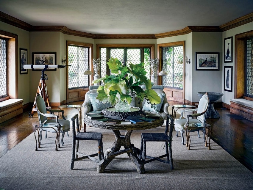 Elegant living room with large windows, eclectic furniture, a telescope, and a central table with a leafy plant centerpiece.