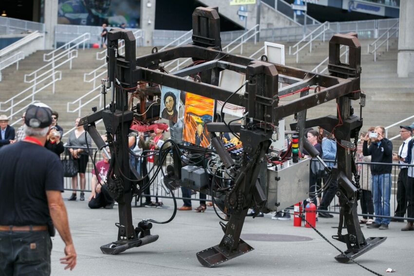 Walking robotic art display with electronic components in an outdoor setting, surrounded by a crowd of onlookers.