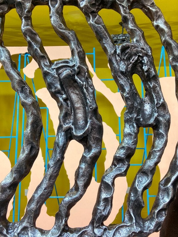 Abstract metal sculpture with twisted, coiled forms against a vibrant yellow and blue background.