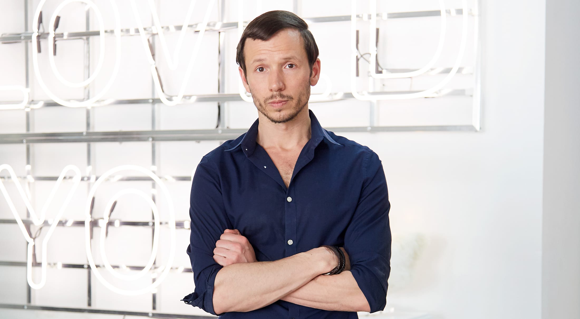 Man with crossed arms, wearing a navy shirt, standing in front of neon sign with blurred letters in a modern setting.