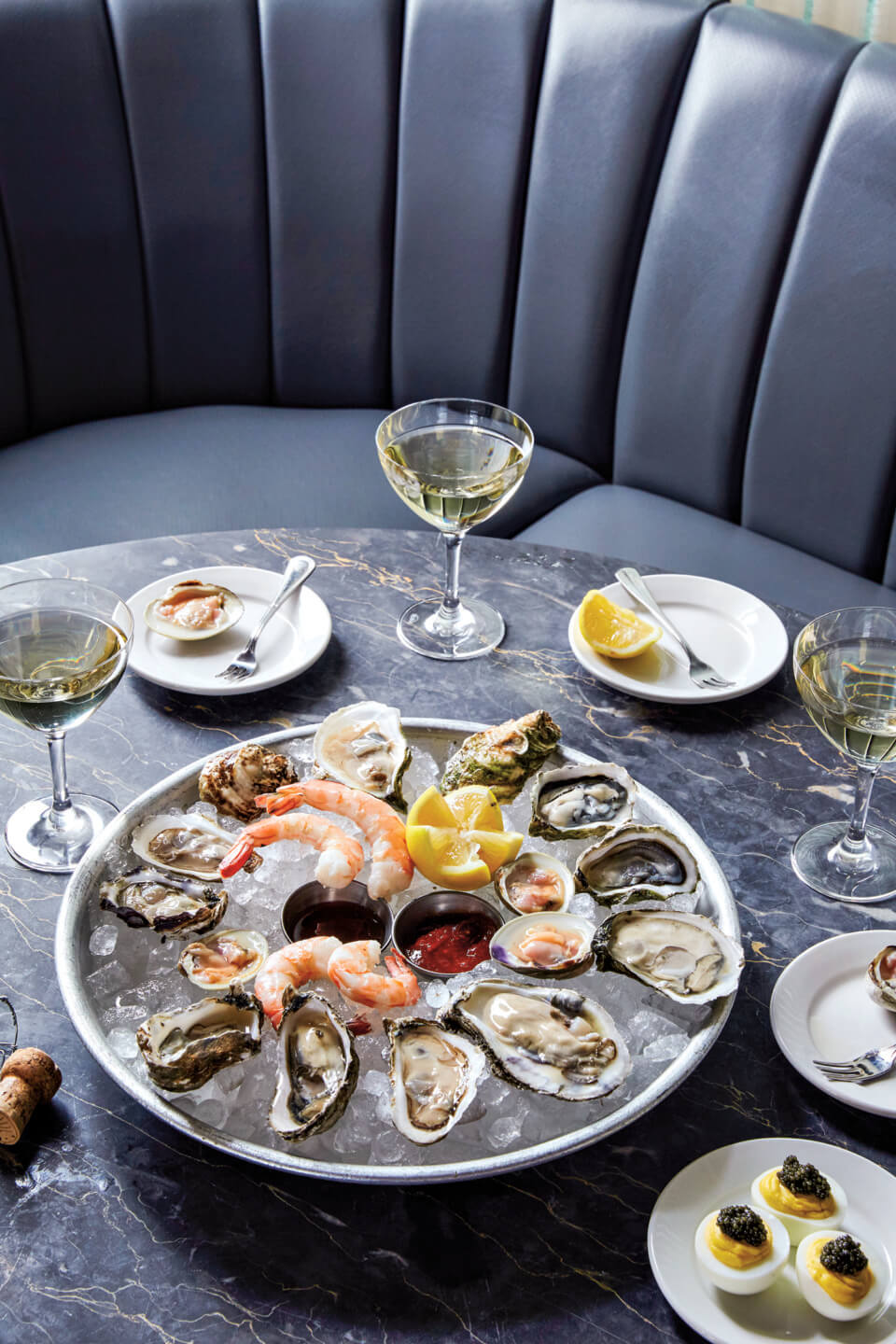 Seafood platter with oysters, shrimp, and lemon wedges on ice, served with glasses of white wine.
