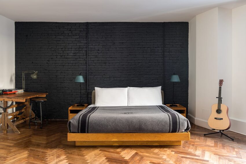 Cozy bedroom with a wooden bed, black accent wall, desk, industrial lamps, and a guitar on herringbone wood floor.