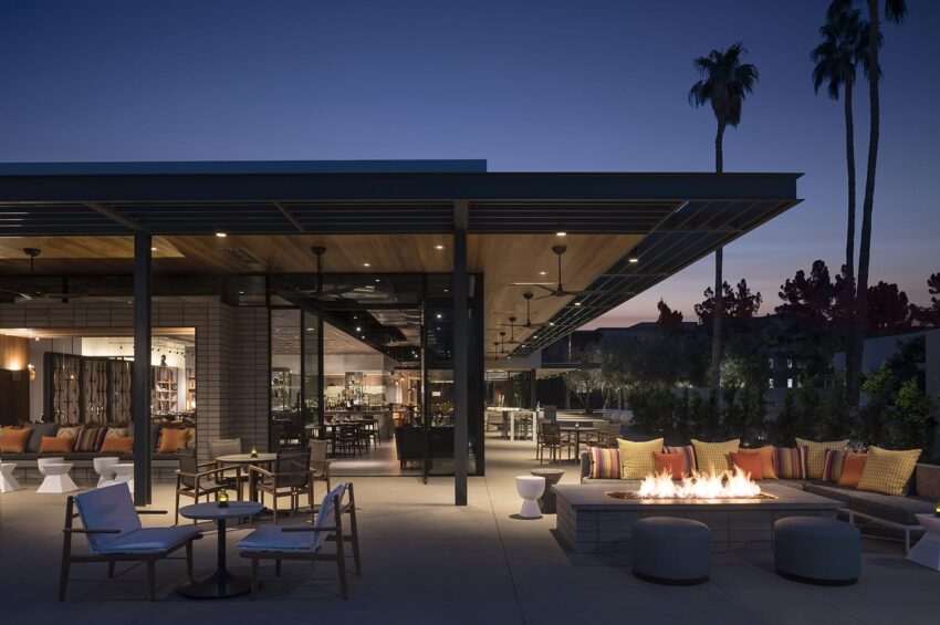 Outdoor patio with modern furniture, fire pit, and palm trees at dusk, with indoor area visible through large glass walls.