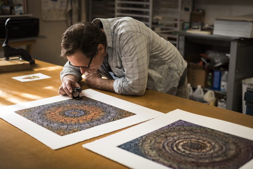 Man examining detailed artwork with a magnifying glass in a workshop, surrounded by creative materials and papers.