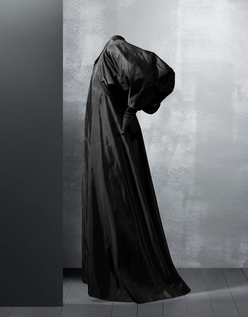 A person in a flowing, oversized black garment stands against a textured gray wall with their head bent forward.
