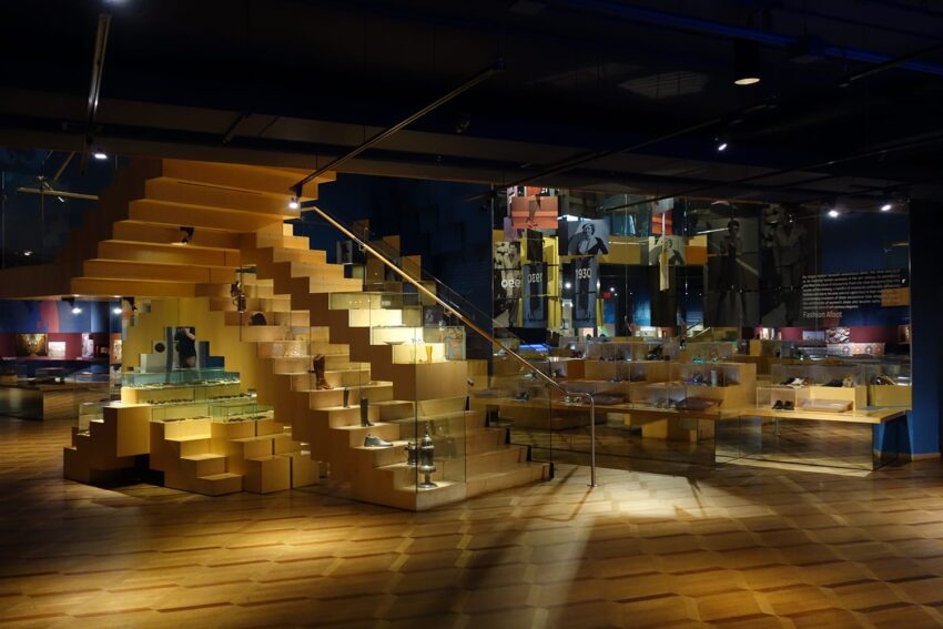 Modern museum exhibit with wooden staircase, reflective surfaces, and display cases under soft lighting.