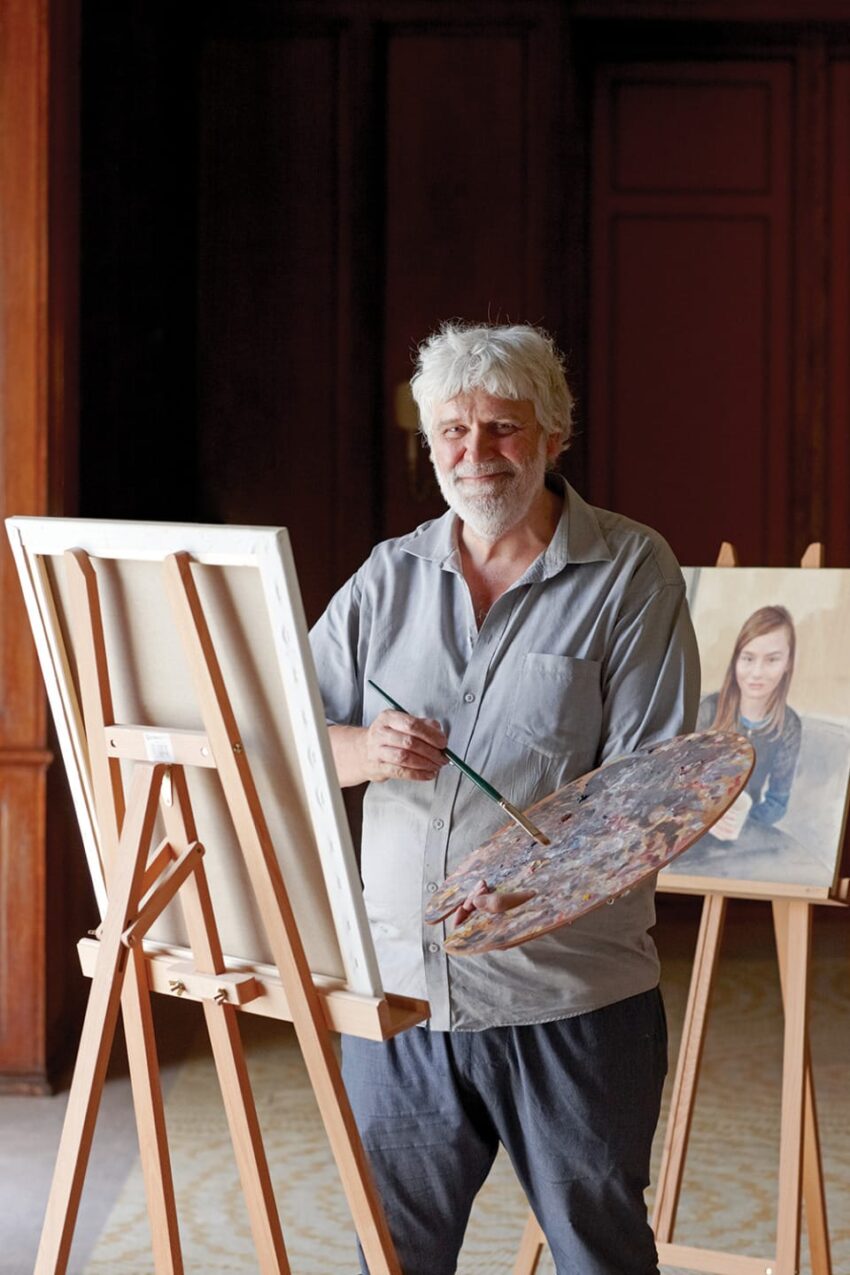 Artist standing in a studio holding a paintbrush and palette, with a canvas and a portrait of a young person behind him.