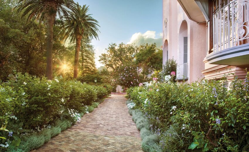 Garden path lined with colorful flowers and lush greenery beside a peach-toned building, with the sun setting behind palm trees.