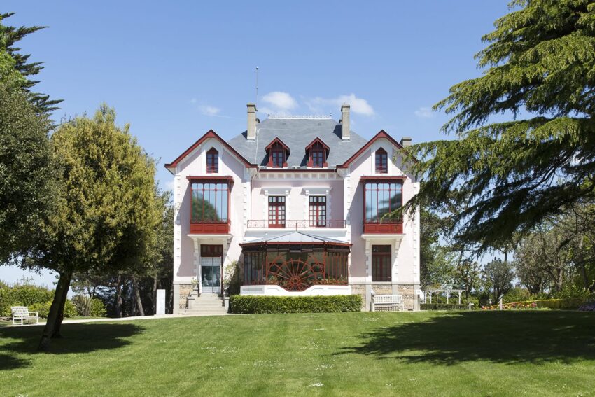 Large white mansion with red accents, surrounded by a lush green lawn and tall trees, under a clear blue sky.