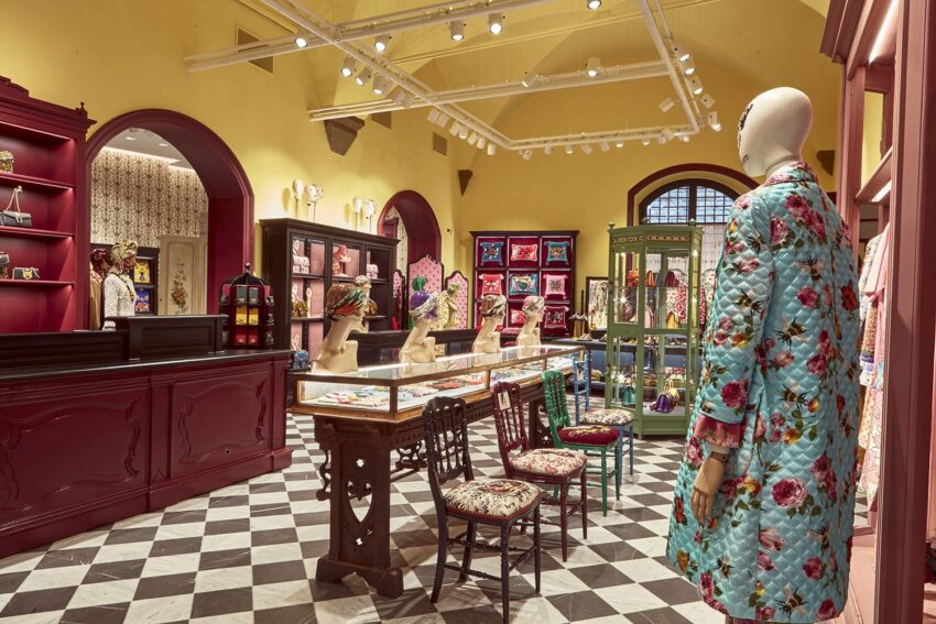 Colorful boutique interior with floral patterns, mannequin in a floral coat, ornate chairs, and glass display cases.