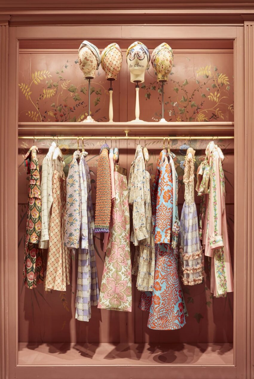 Vintage-style dresses hanging on a rack with decorative headpieces above against a floral painted wall.